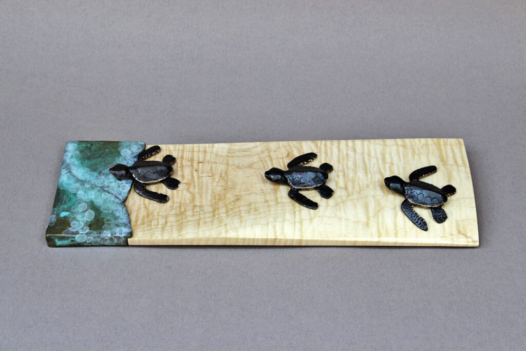 Three baby sea turtles heading out to sea. The turtles and the surf are bronze, and the sand is represented by a piece of curly maple.