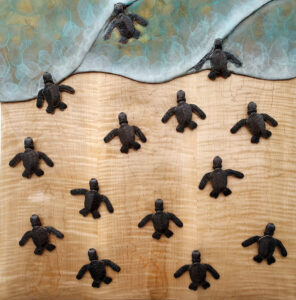 A bronze sculpture depicts three baby sea turtles making their way towards the ocean. The turtles and the surf are crafted in bronze, while the sand is represented by a beautiful piece of curly maple with a swirling grain pattern that resembles the ripples and waves of the sea.